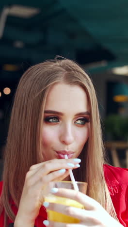 gorgeous woman drinking juice. beautiful young lady in red dress sitting on a swing and drinking cold beverage. attractive model relaxing in a cafe. vertical video