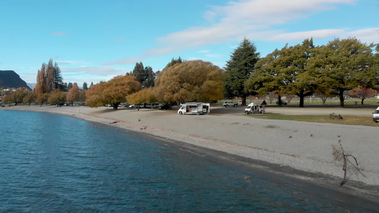 가을에 뉴질랜드 와나카 호수에 있는 아름다운 푸른 호수 위의 해변에 있는 캠퍼밴을 향해 날아가는 slowmo 공중 드론