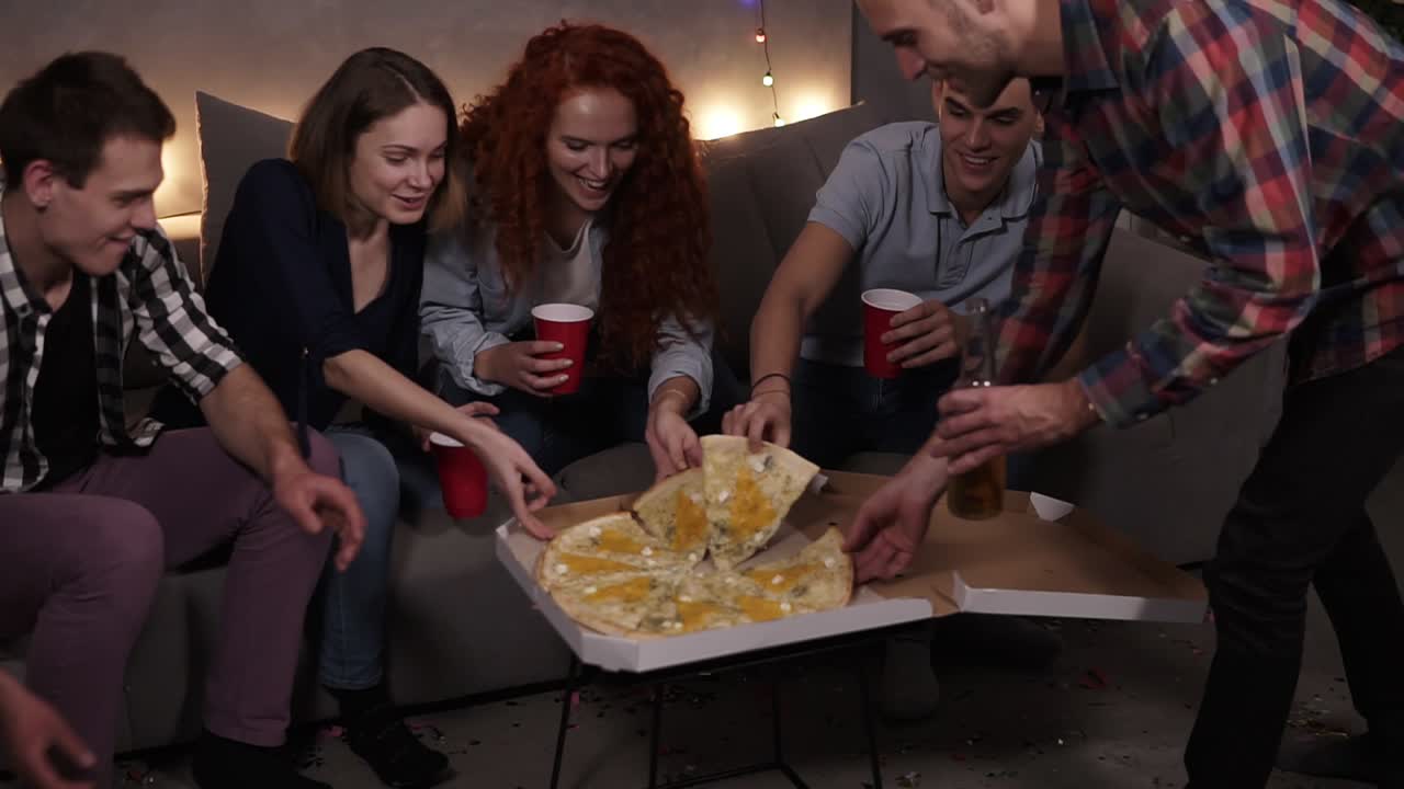 Diverse people, friends got together to eat pizza at home. Got delivery Italian pizza with cheese, taking slices, holding red cups and bottles. Starting home pizza party. Garlands light on the background