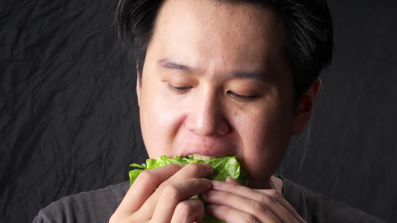 暗い背景をバックに、新鮮なレタスの葉を食べるアジア人男性。健康的な食生活のコンセプトを表している。