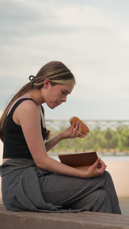woman wearing black tank top seated outdoors, eating burger while reading book with a decorative fence and bright sky in the background, reading and snacking in an open-air setting