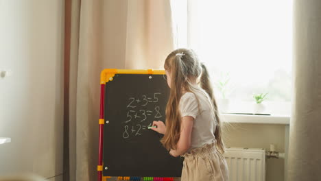 girl writes answer on blackboard younger child shows digit