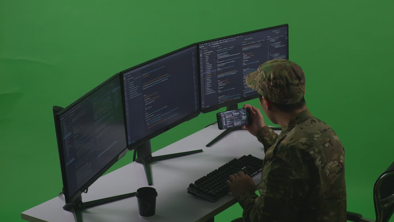 Side View Of Happy Asian Man Soldier Developer Looking At Code On Mobile Phone While Write Code With Multiple Computer Screens In Green Screen Studio