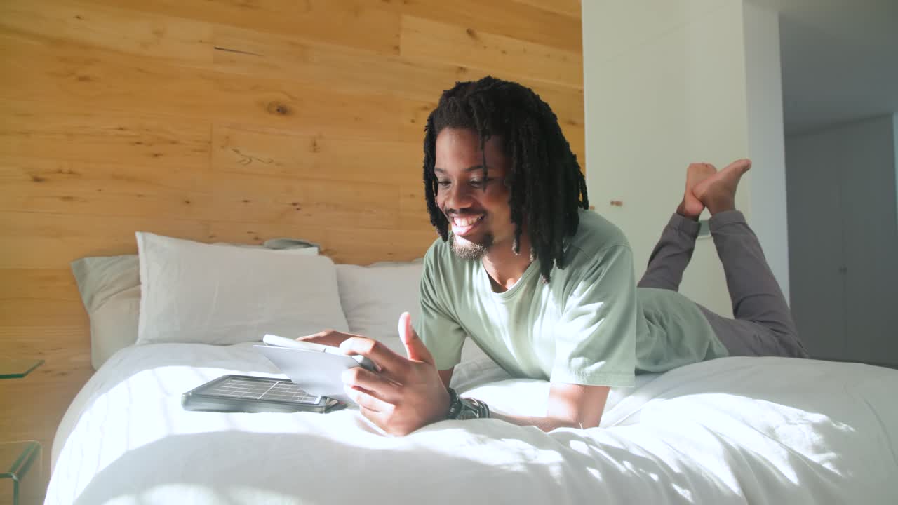 African American man entering bedroom, dropping onto bed, reaching tablet, reading paper, smiling