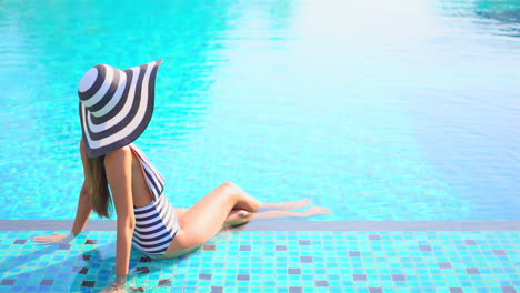 Vista-Trasera-De-Una-Turista-Sentada-Cerca-De-Una-Piscina,-Usando-Traje-De-Baño-Blanco-Y-Negro-Y-Un-Gran-Sombrero-Flexible,-Empapando-Sus-Pies-En-La-Piscina-Al-Aire-Libre