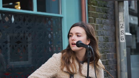 Female-Musician-Busking-Playing-Acoustic-Guitar-And-Singing-To-Crowd-Outdoors-In-Street