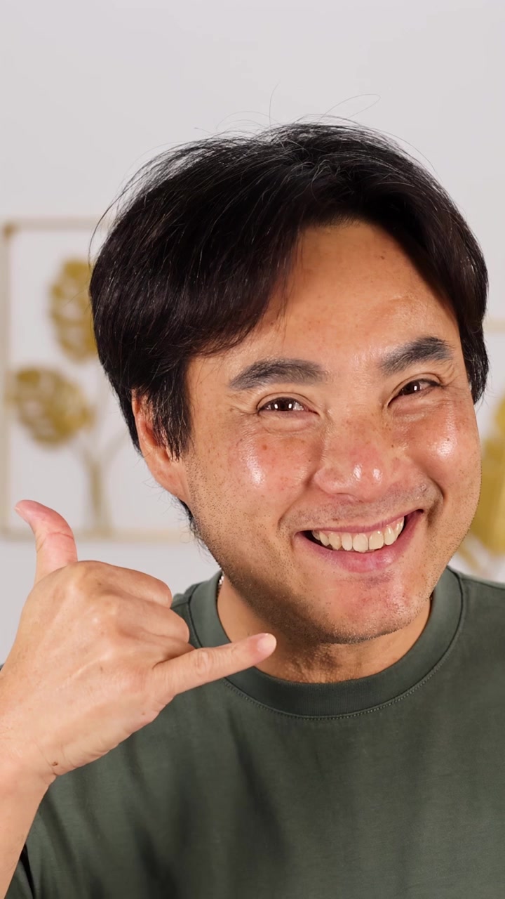 Uomo asiatico sorridente con una camicia verde fa il gesto di 'chiamami' al chiuso, amichevole e invitante.