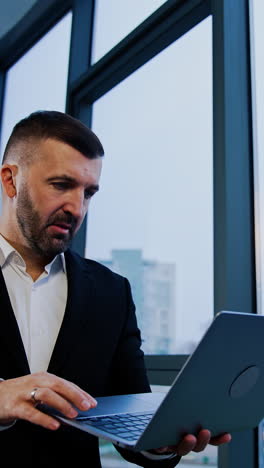 thoughtful businessman stands holding laptop in hands. male boss types on computer waving head disapprovingly. low angle view. vertical video