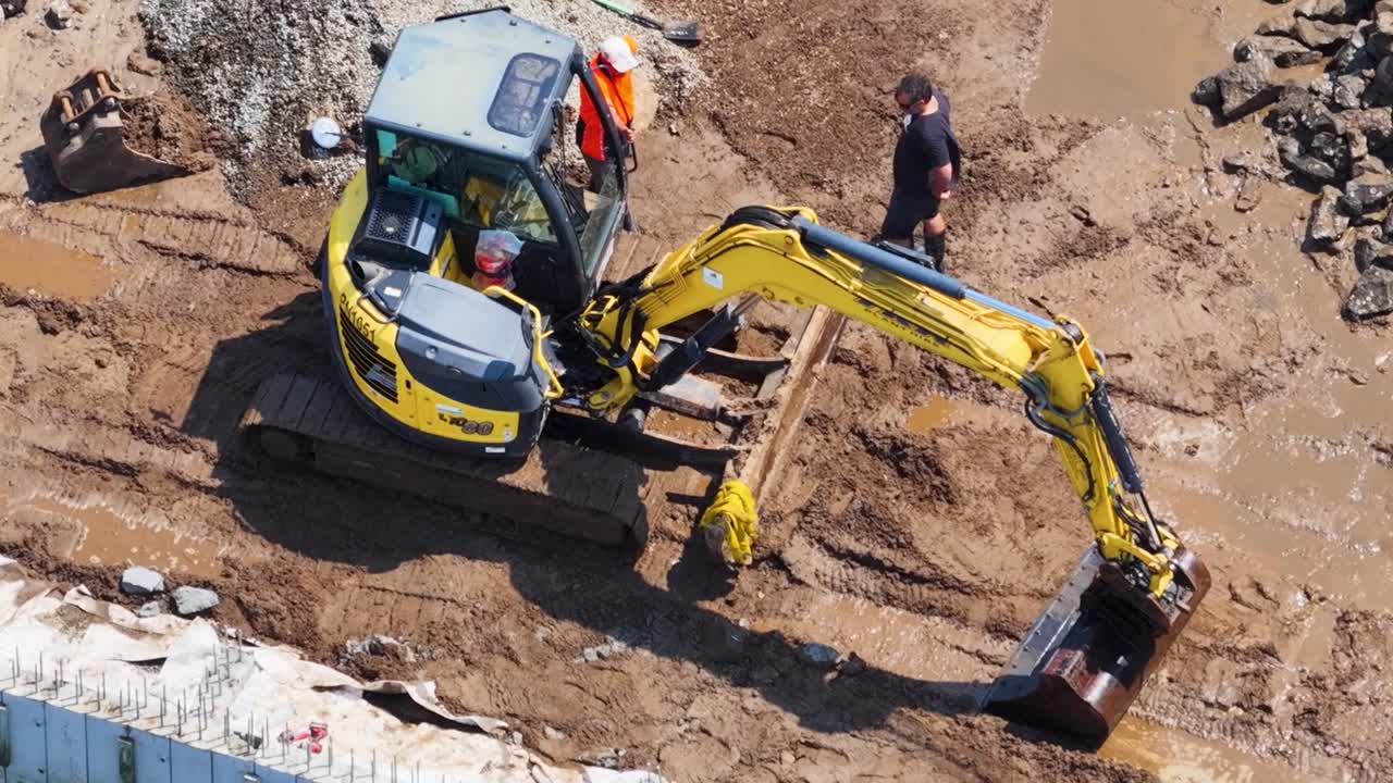 Luftfoto af en kompakt gravemaskine og arbejdere, der forbereder et mudret fundament på en byggeplads