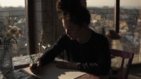 young cute female artist with curly hair pulled in a bun, sitting by the table and drawing with a pen fully concentrated on the process of creation. art craft, inspiration.