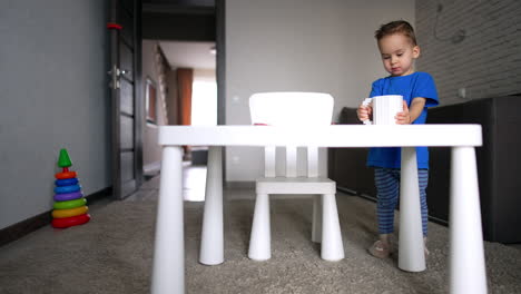 little male kid stands at the desk holding a big blue cup. lovely boy pretends drinking from a cup.
