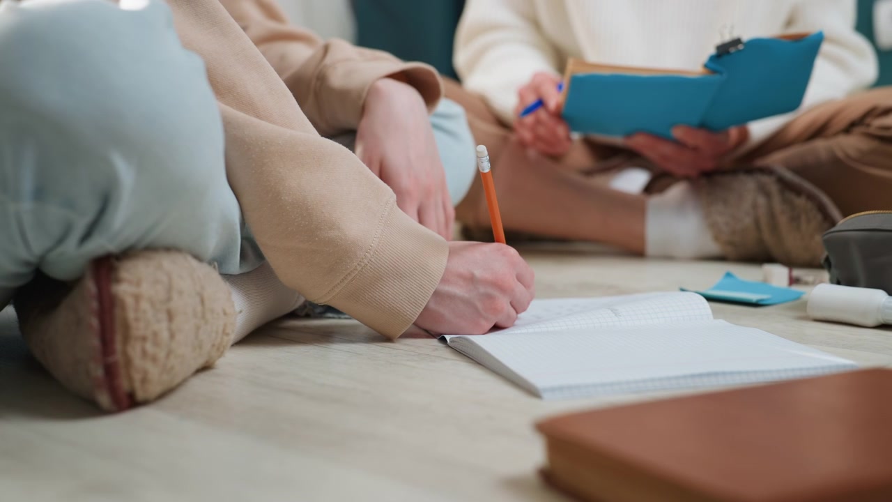 Sessione di tutoraggio a casa accogliente, tutor che guida esercizi di grammatica sul pavimento, studente che scrive su un quaderno con una matita, appunti su una cartella blu, abbigliamento casual rilassato, illuminazione soffusa, focus sul mentoring di supporto