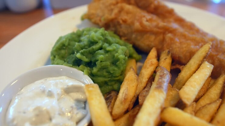 Fish n chips with golden fries, crispy battered fish, and a serving of mushy peas is sitting on a table, with a side of tartar sauce