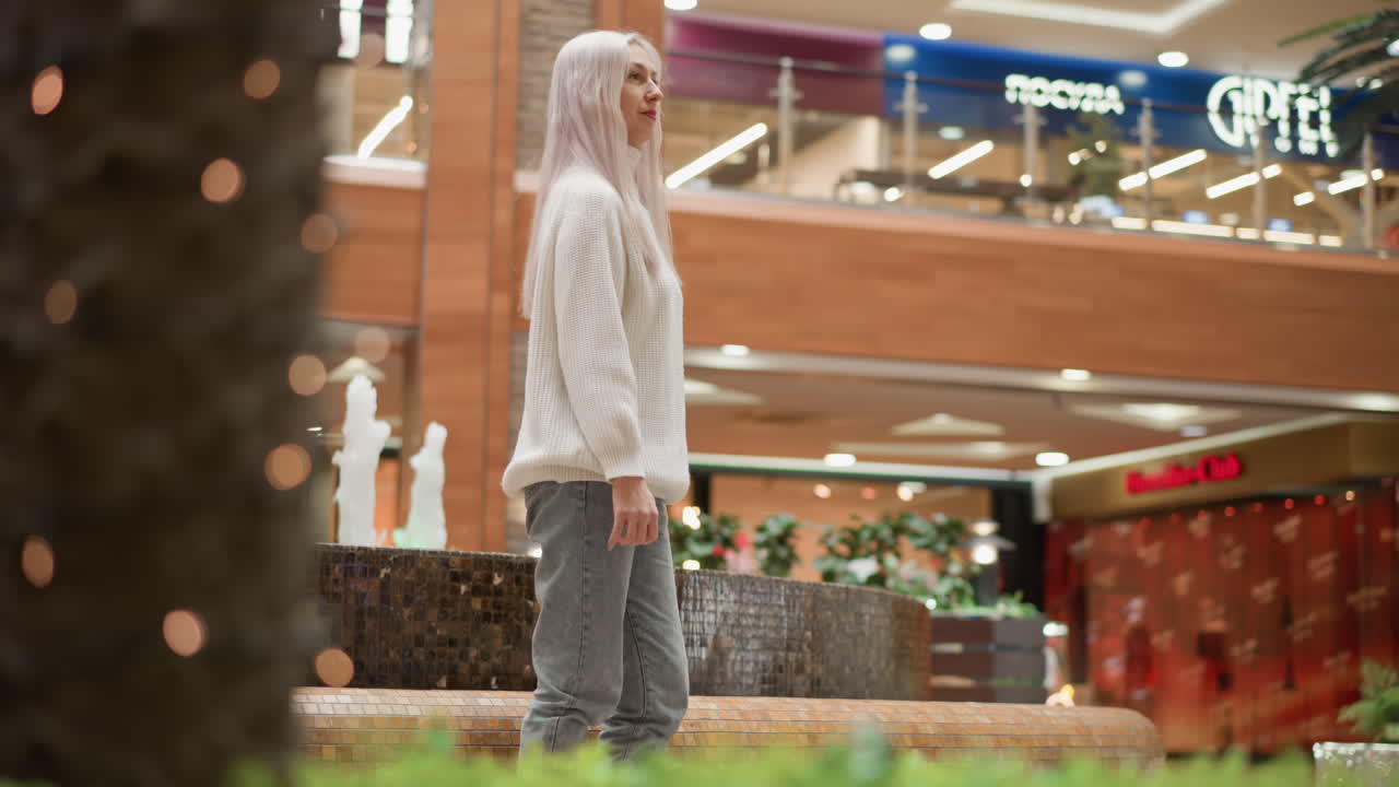 Reflective woman sits by indoor fountain in mall, gracefully turning to watch flowing water, ambient shoppers walking past under gallery lighting, mix of wood and tile surfaces, modern retail
