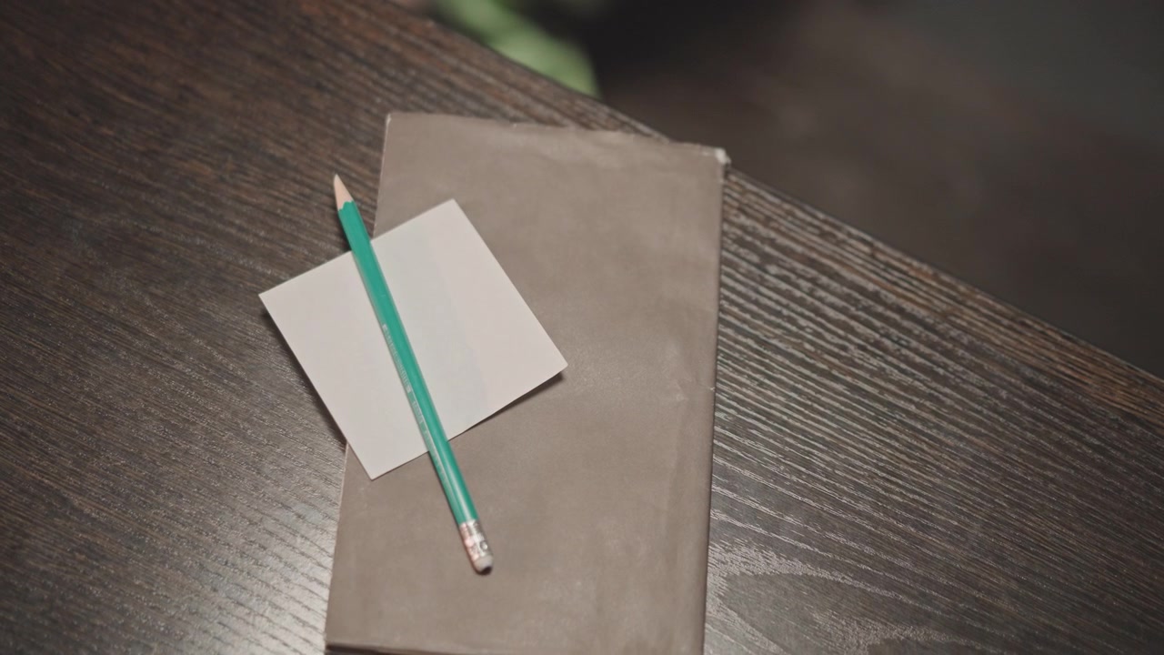 Pencil on notebook atop wooden desk, music teacher prepping lesson notes with green pencil and small notecard, soft warm lighting and shallow depth of field, overhead closeup showing fabric sleeve