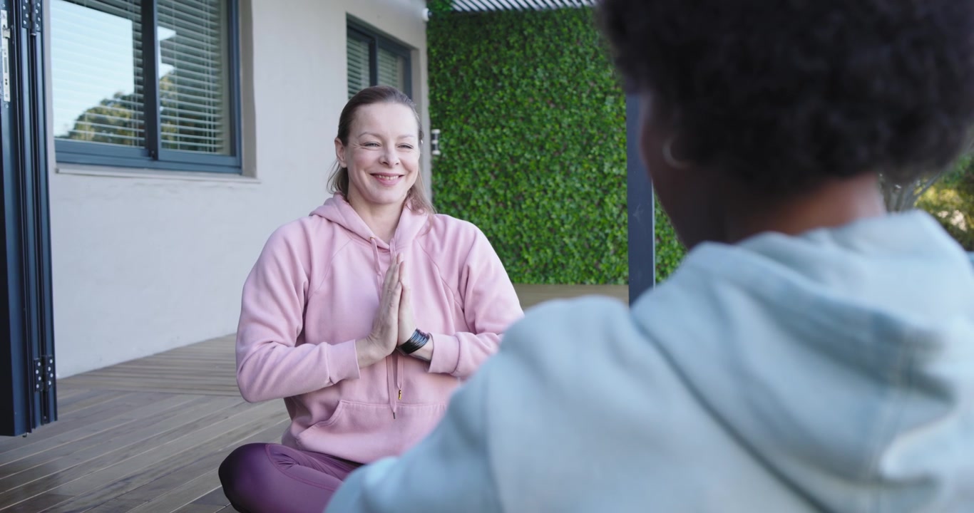 Diverse women meditating inhaling lifting arms to prayer on wood deck checking smartwatch for calm