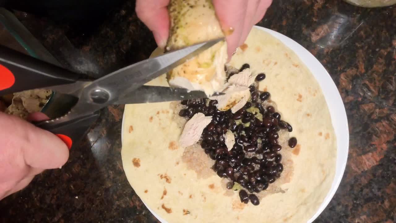 Close-up top-view footage captures a chef cutting cooked chicken with scissors to prepare a fresh tortilla wrap in a kitchen