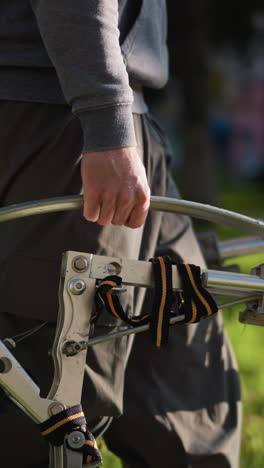 persona con pantalones de color gris oscuro y sudadera a juego agarra una barra metálica curva, posiblemente parte de un dispositivo para caminar o pilotes, al aire libre en hierba verde borrosa durante el día; bajo ángulo de la cámara captura
