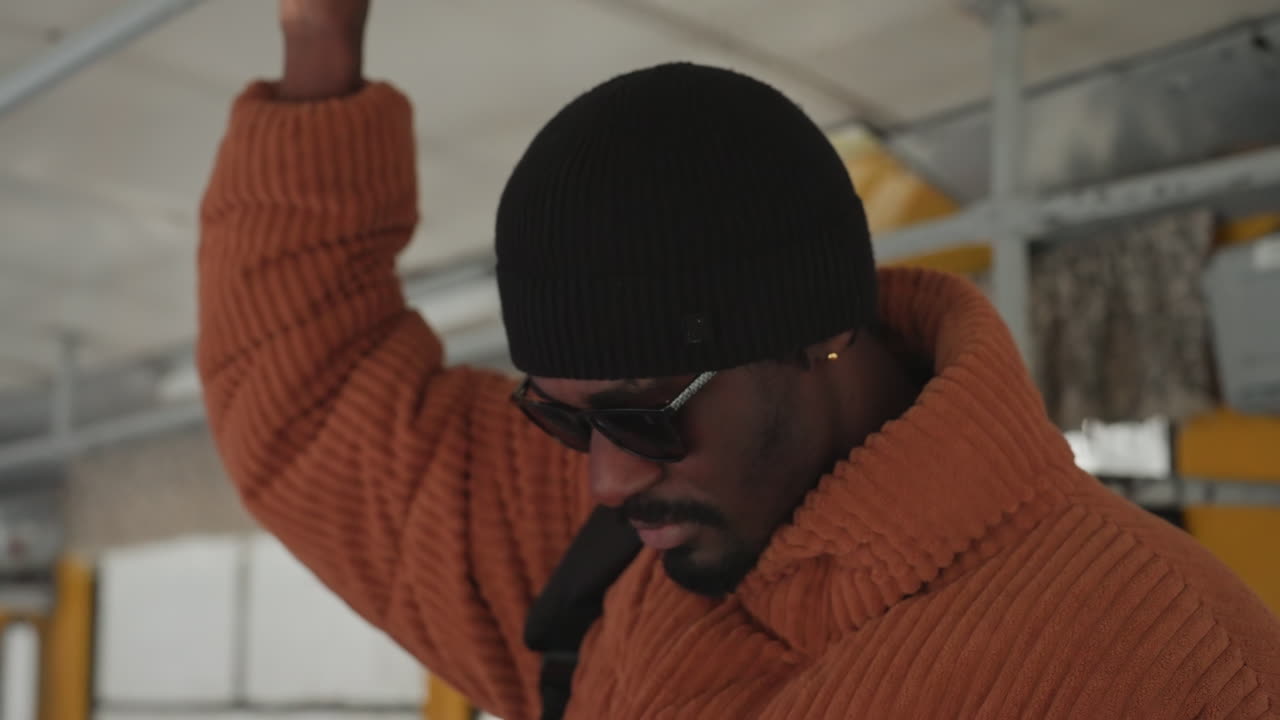 Man holding bus rail wearing orange knit coat, black beanie, sunglasses backpack over shoulder, looking out window, soft diffused light, candid city commute freelance photographer heading