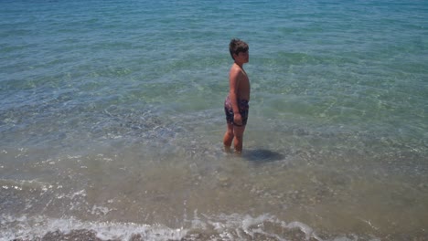 Wide-tilt-down-and-up-establishing-shot-of-caucasian-10-years-old-boy,-entering-the-clear-water-of-sea-at-Kalamata,-Peloponnese,,-Greece-4K