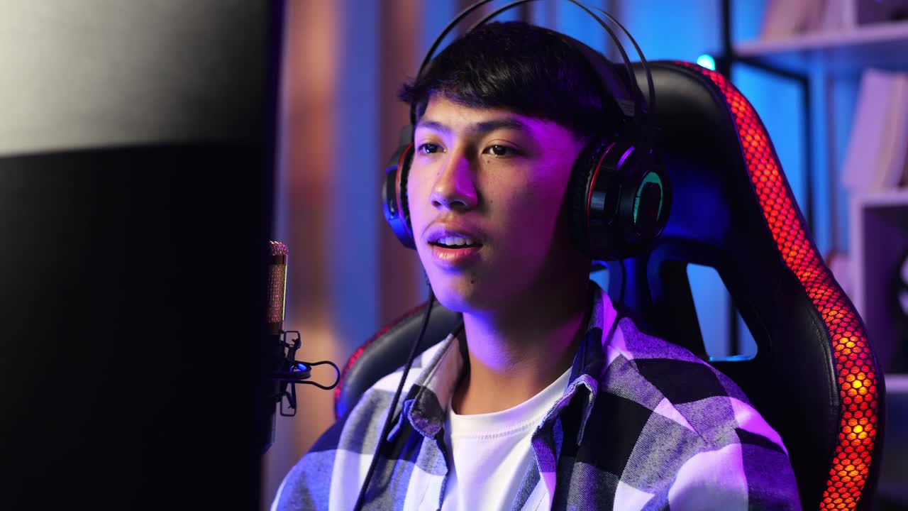 primer plano de un niño asiático sonriendo a la cámara mientras juega un juego a través de la red en una computadora personal. videojuego de transmisión en vivo, escritorio iluminado por luz de tira led rgb