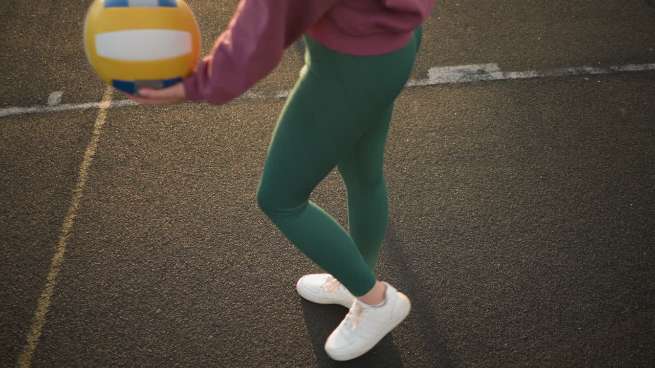 vista hacia abajo de la atleta en leggings verdes y zapatos blancos rebotando la pelota de voleibol antes de servirla mientras salta, capturando el movimiento dinámico y la energía durante la práctica de voleibal