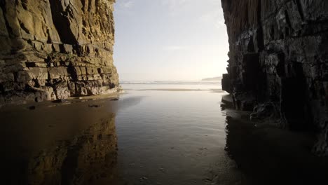 a huge sea cave with tide water coming through during sunset by the ocean with light reflected on the water above the sand