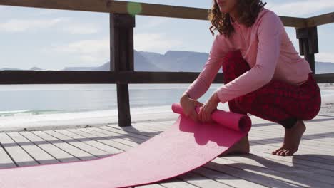 healthy mixed race woman practicing yoga outdoors, squatting and rolling up mat by the sea