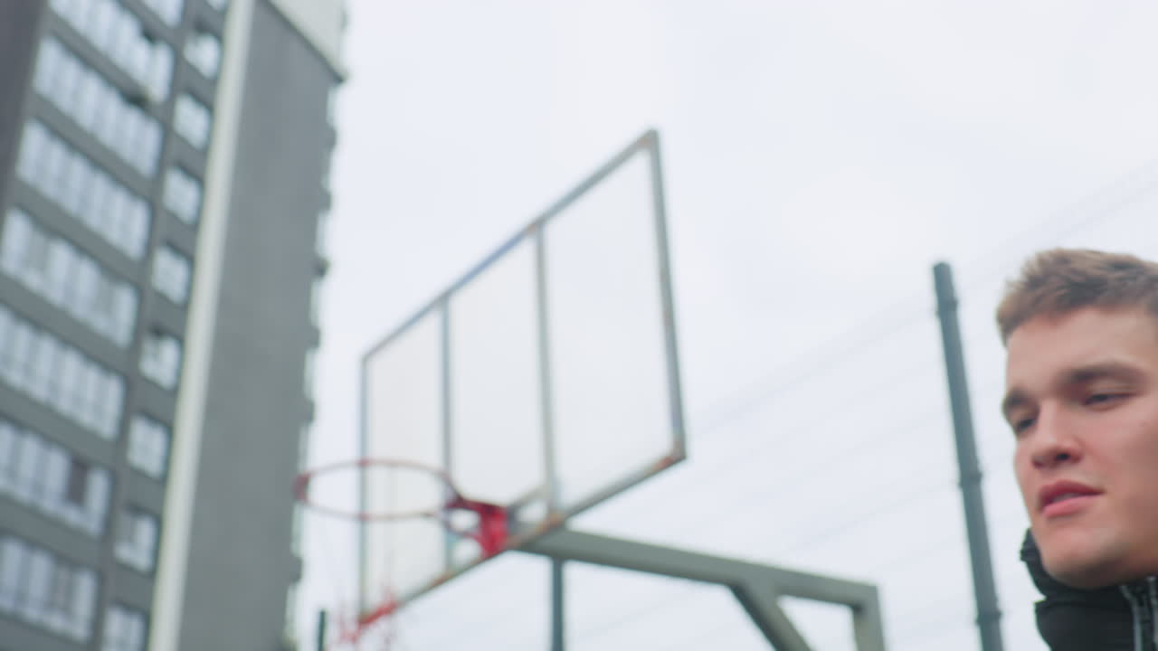 Giovane bianco fa roteare un pallone da basket su un dito, campo urbano all'aperto con grattacielo e canestro, primo piano sul cerchio e sul pallone texturizzato, espressione sicura di sé in giacca streetwear, atmosfera grintosa e giocosa