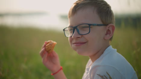 primer plano de un niño con gafas y una camisa blanca, sonriendo contento mientras sostiene un pastel de perrito caliente en un campo cubierto de hierba. la expresión feliz del niño refleja un momento de disfrute pacífico