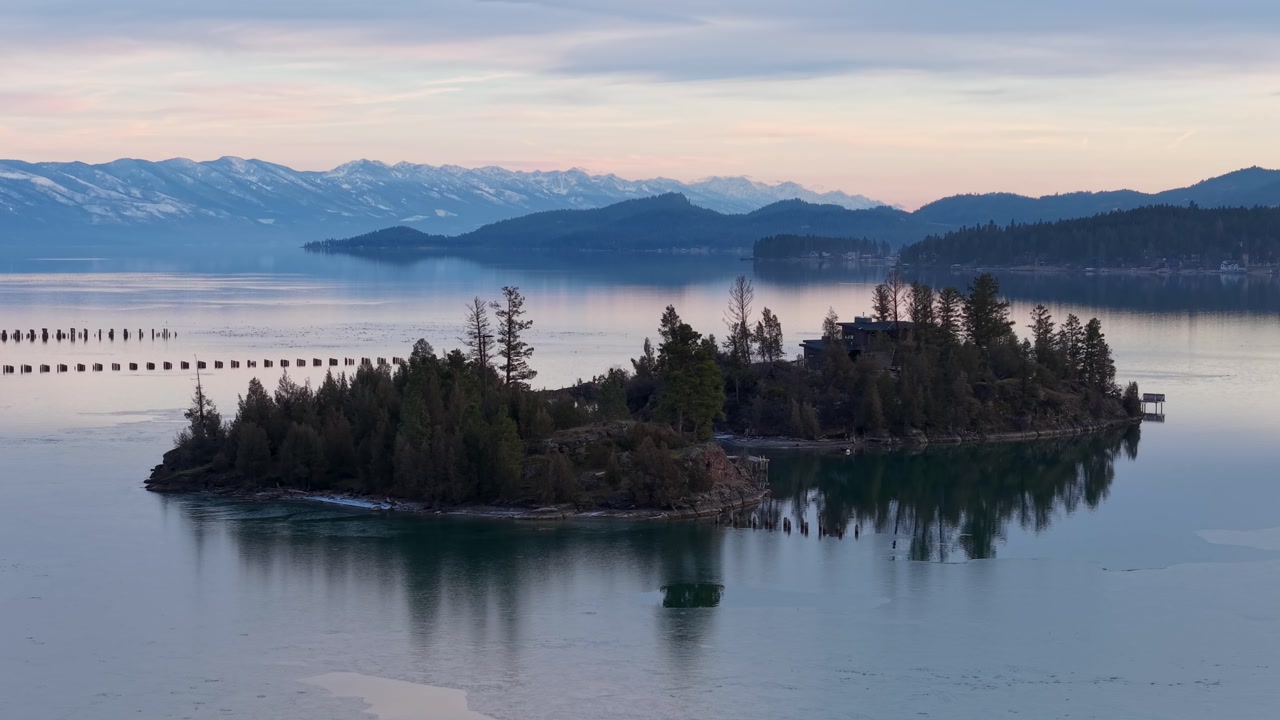 Drone aerial view of a tree-covered island on a calm lake with snow-capped mountains and cloudy sky at dusk.