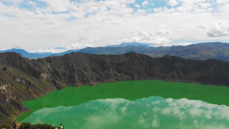 aerial drone shot reveals the turquoise crater of quilotoa lake with tilt down view and tourists gather along the crater's edge in awe