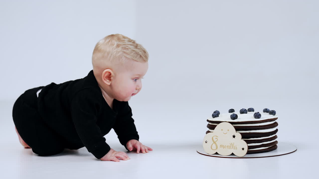 Lovely cute blond baby getting closer to the cake standing in front of him. Child near the tasty dessert. White backdrop.