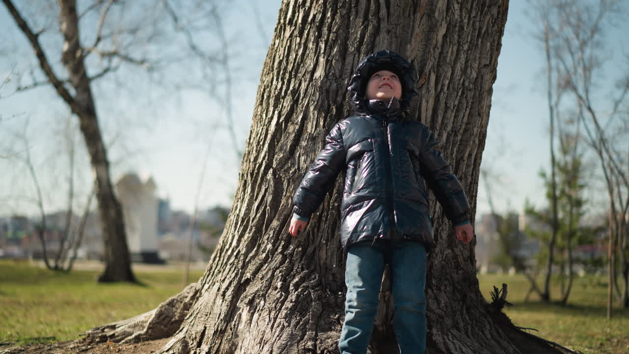 A young boy leans his back against a large tree trunk, looking up with a smile, holding small stones in both hands, he is dressed in a shiny black jacket, jeans, and boots, with trees around