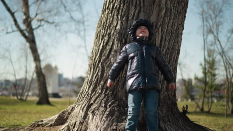 a young boy leans his back against a large tree trunk, looking up with a smile, holding small stones in both hands, he is dressed in a shiny black jacket, jeans, and boots, with trees around