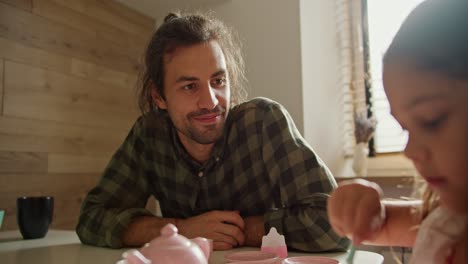 Over-the-shoulder-little-brunette-girl-in-a-pink-dress-plays-tea-party-with-the-help-of-toy-pink-cups-and-teapots-together-with-her-happy-brunette-man-father-in-a-checkered-green-shirt-in-a-modern-kitchen