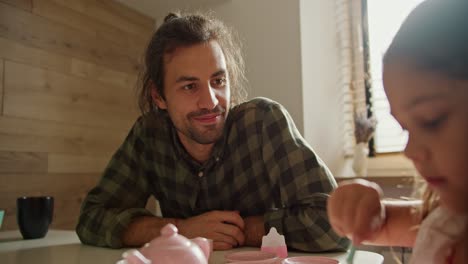 over the shoulder little brunette girl in a pink dress plays tea party with the help of toy pink cups and teapots together with her happy brunette man father in a checkered green shirt in a modern kitchen