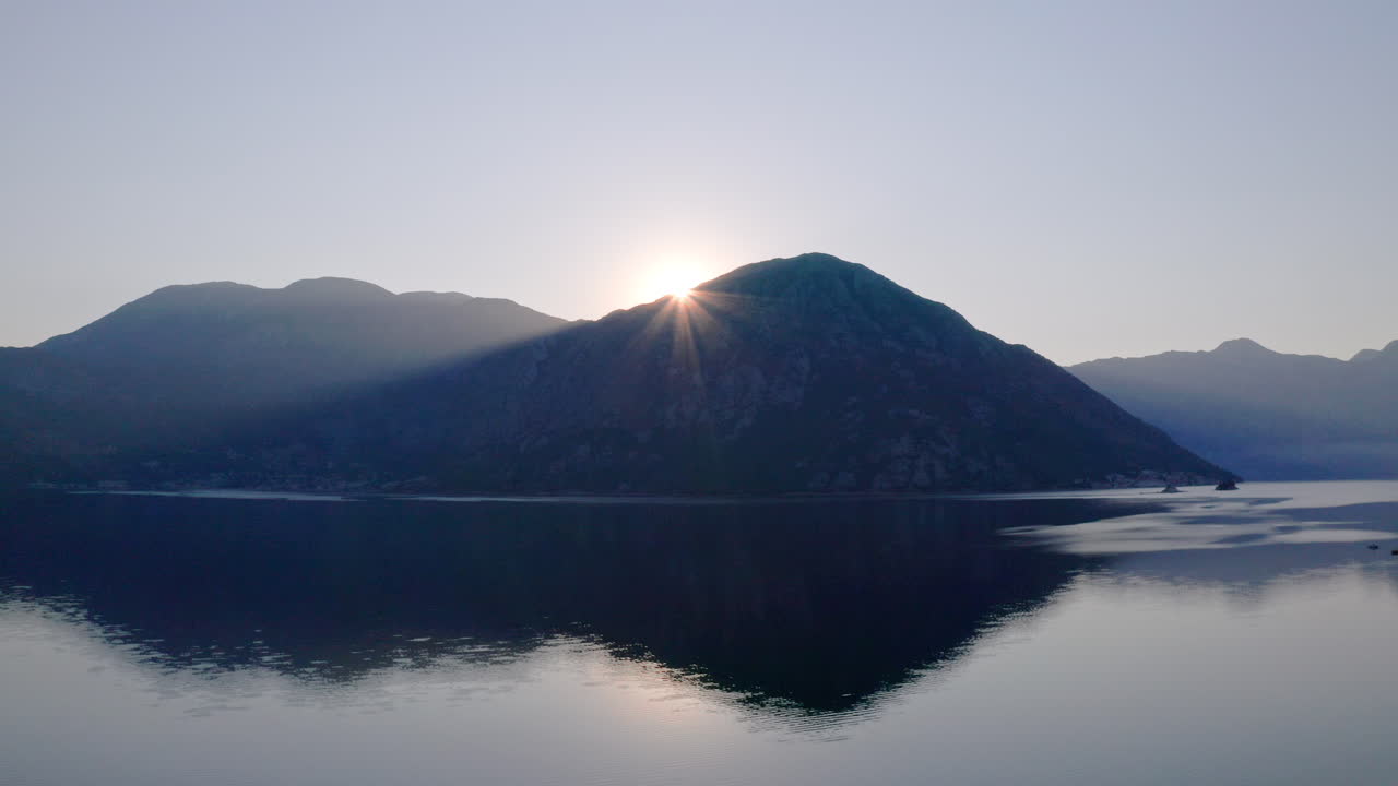 Zoomaava ilmakuva Kotorinlahdesta Montenegrossa, kun aurinko nousee ympäröivien vuorten takaa ja valaisee hitaasti maiseman. Auringonnousu merenlahden ja ympäröivien vuorten yllä.