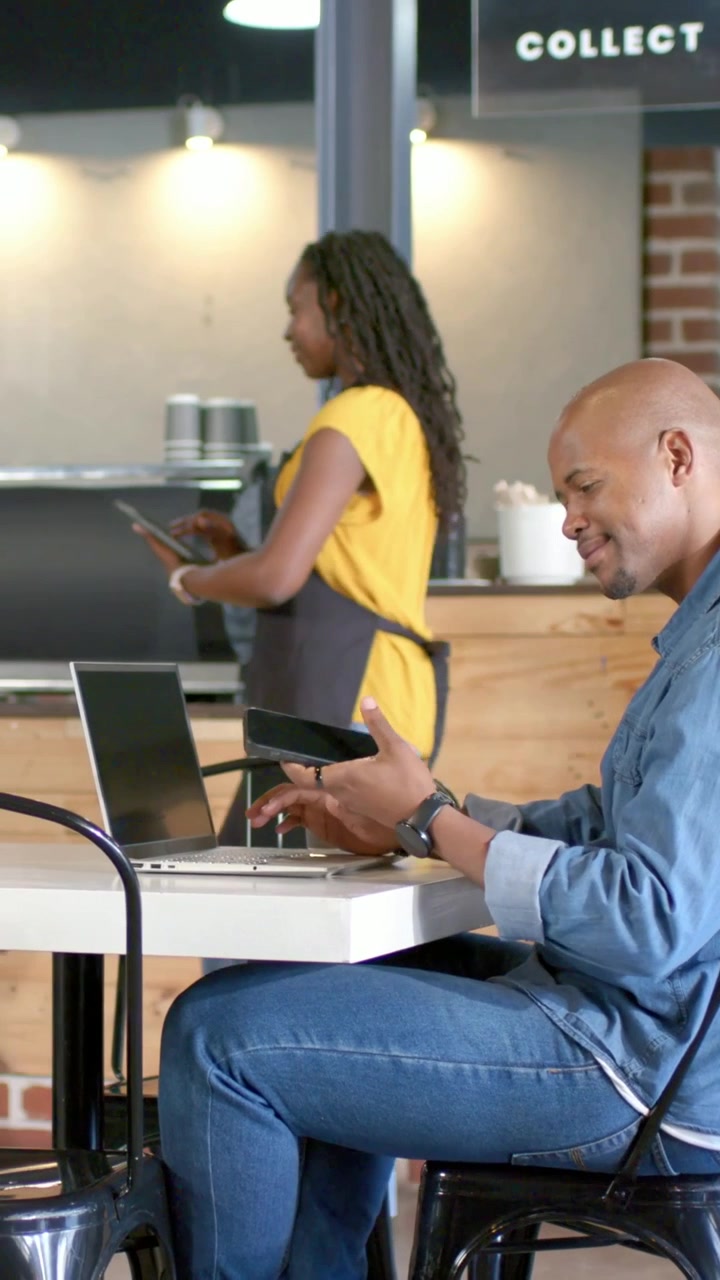 Vídeo vertical: Homem digitando no laptop em um café, barista de avental verificando tablet para pedido no COLLECT