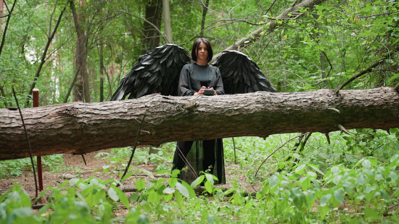 Woman in long black outfit with large dark wings standing behind fallen tree in lush green forest, operating small device with thoughtful expression, surrounded by calm mysterious woodland atmosphere