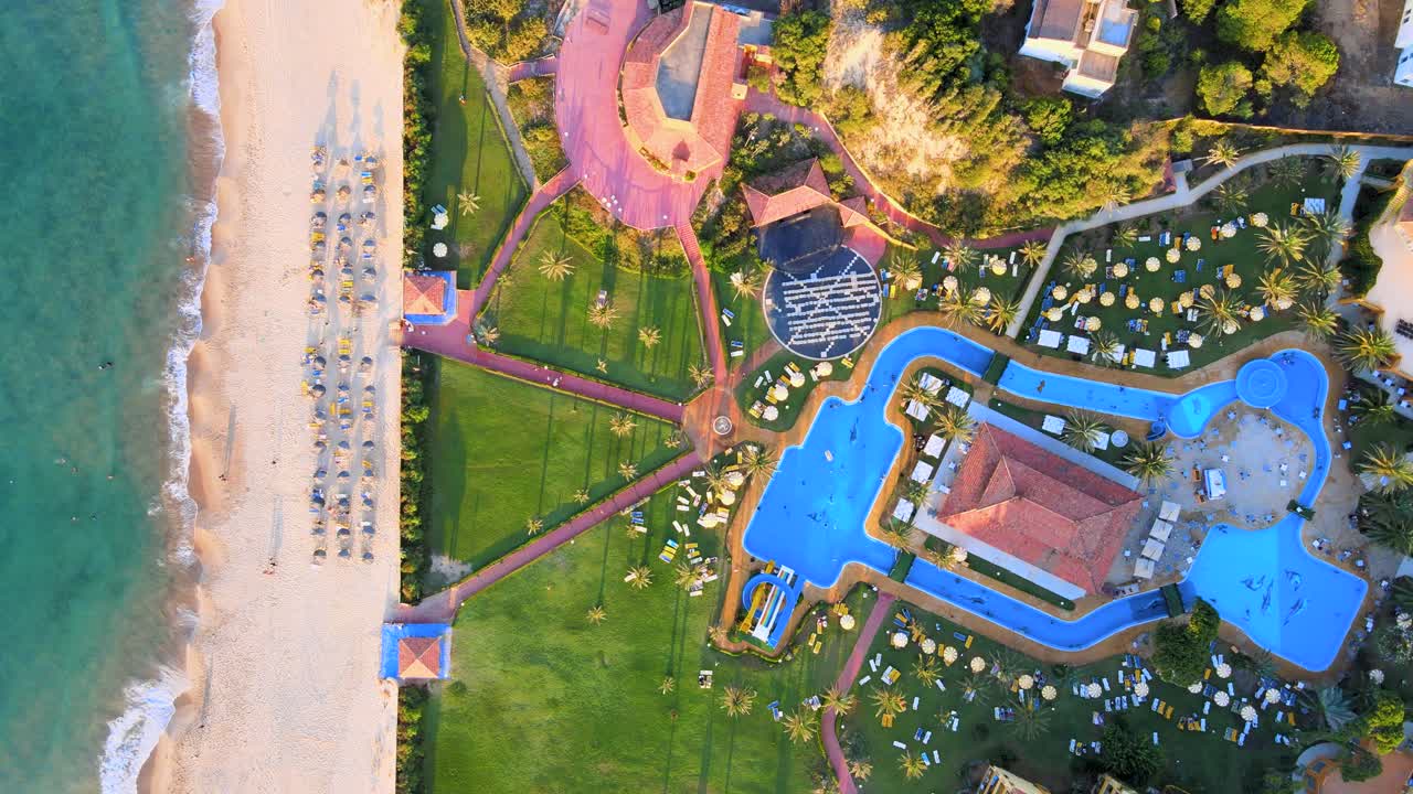 An aerial picture taken over an upscale beach resort, showcasing a beautiful layout that includes blue pools surrounded by green grass and many white umbrellas.