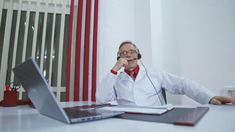Physician with headset talks in medical office. Professional male doctor with headphones in white medical uniform sitting at desk and speaking into microphone in modern hospital office.