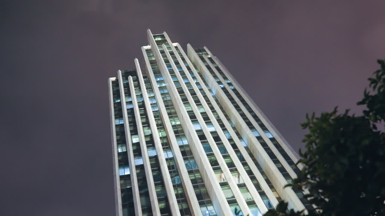 Rascacielos nocturno enmarcado por un primer plano frondoso, fachada de cristal brillante con una cuadrícula de ventanas iluminadas, cielo tormentoso púrpura, una perspectiva ascendente que transmite escala y geometría moderna, urbano sereno pero dramático.