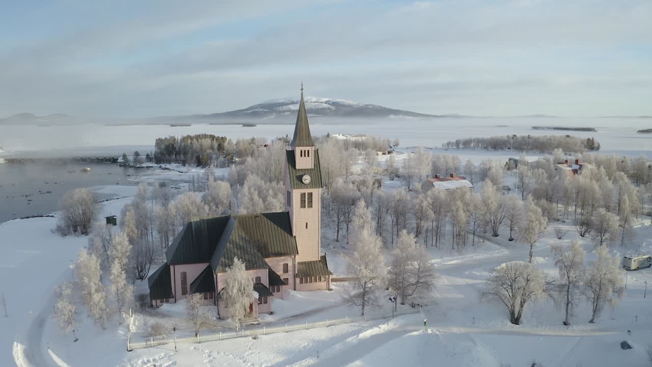 스웨덴 북부의 주변 소나무와 언덕, 얼어붙은 호수를 배경으로 겨울 동안 교회 궤도를 도는 드론 샷