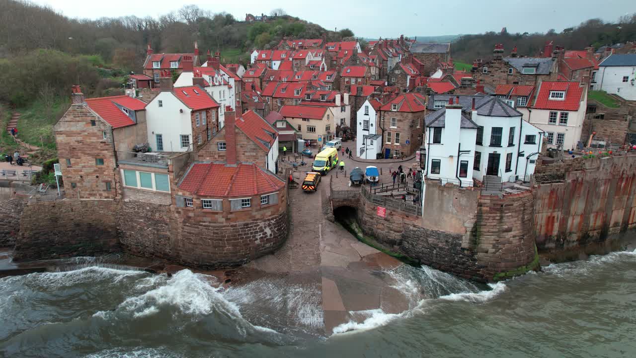 olas rompiendo contra las coloridas casas de la ciudad de robin's hood bay vista aérea estática de cerca