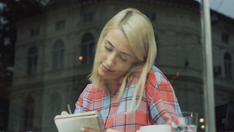 Blonde-Frau-Sitzt-In-Einem-Café-Und-Denkt-Nach,-Während-Sie-Etwas-Mit-Einem-Bleistift-In-Ihr-Notizbuch-Schreibt