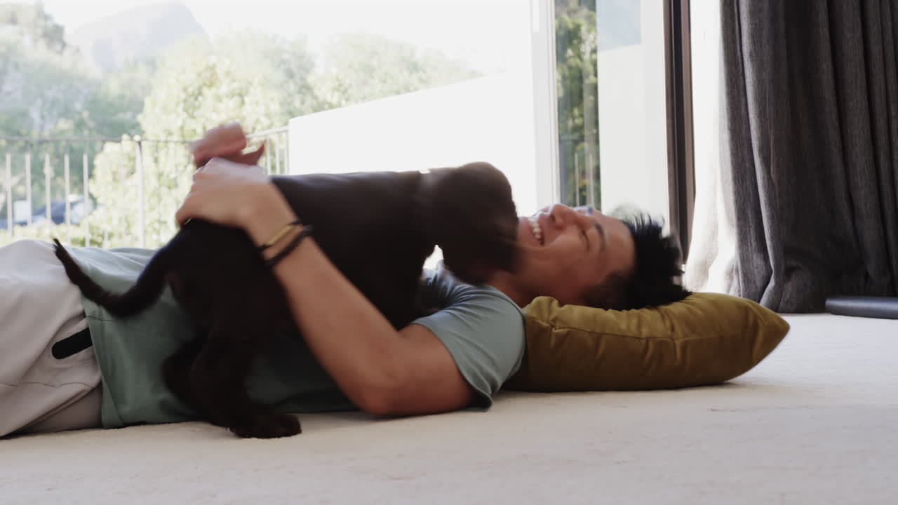 Asian man laughing while playing with dog on floor at home