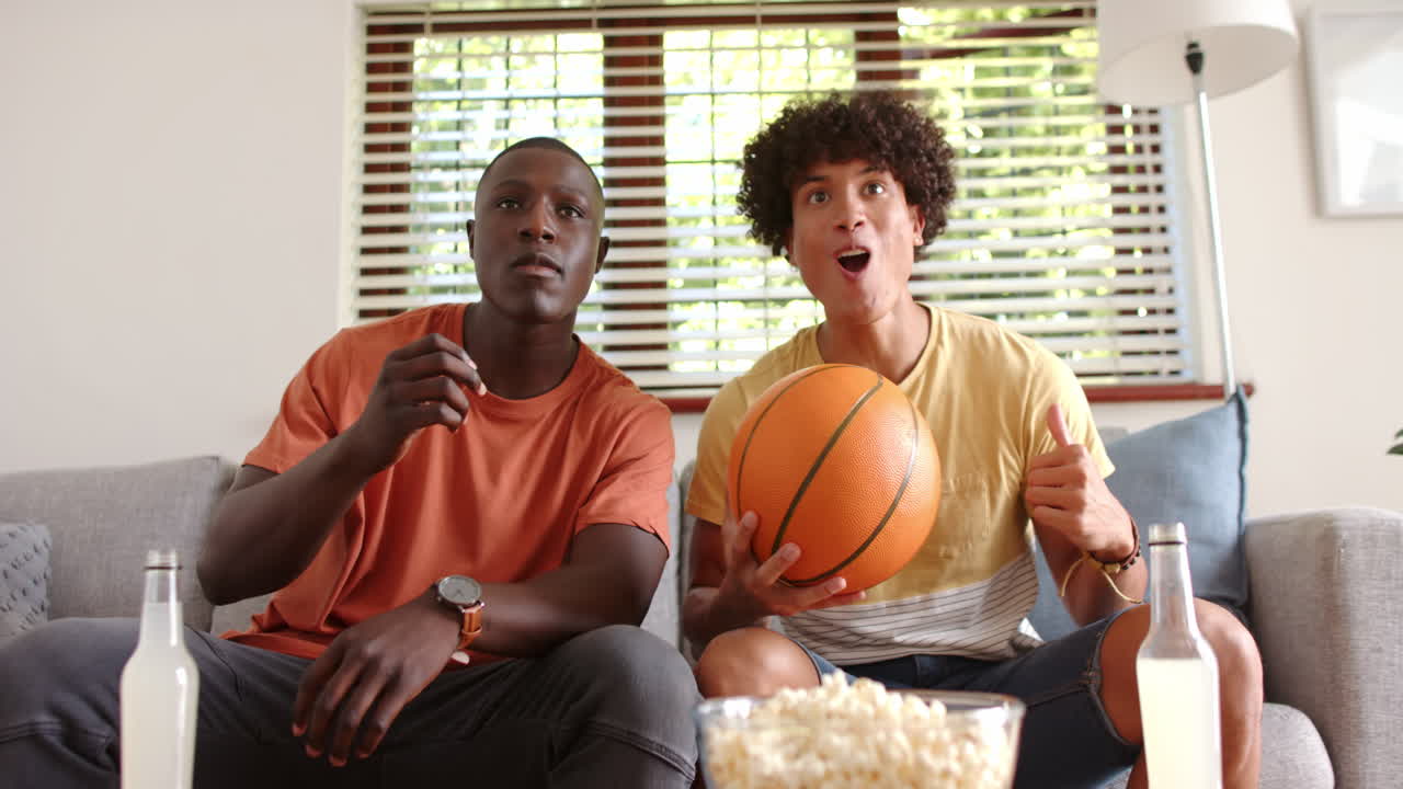 Watching sports game, two excited multiracial male friends holding basketball and sitting, at home