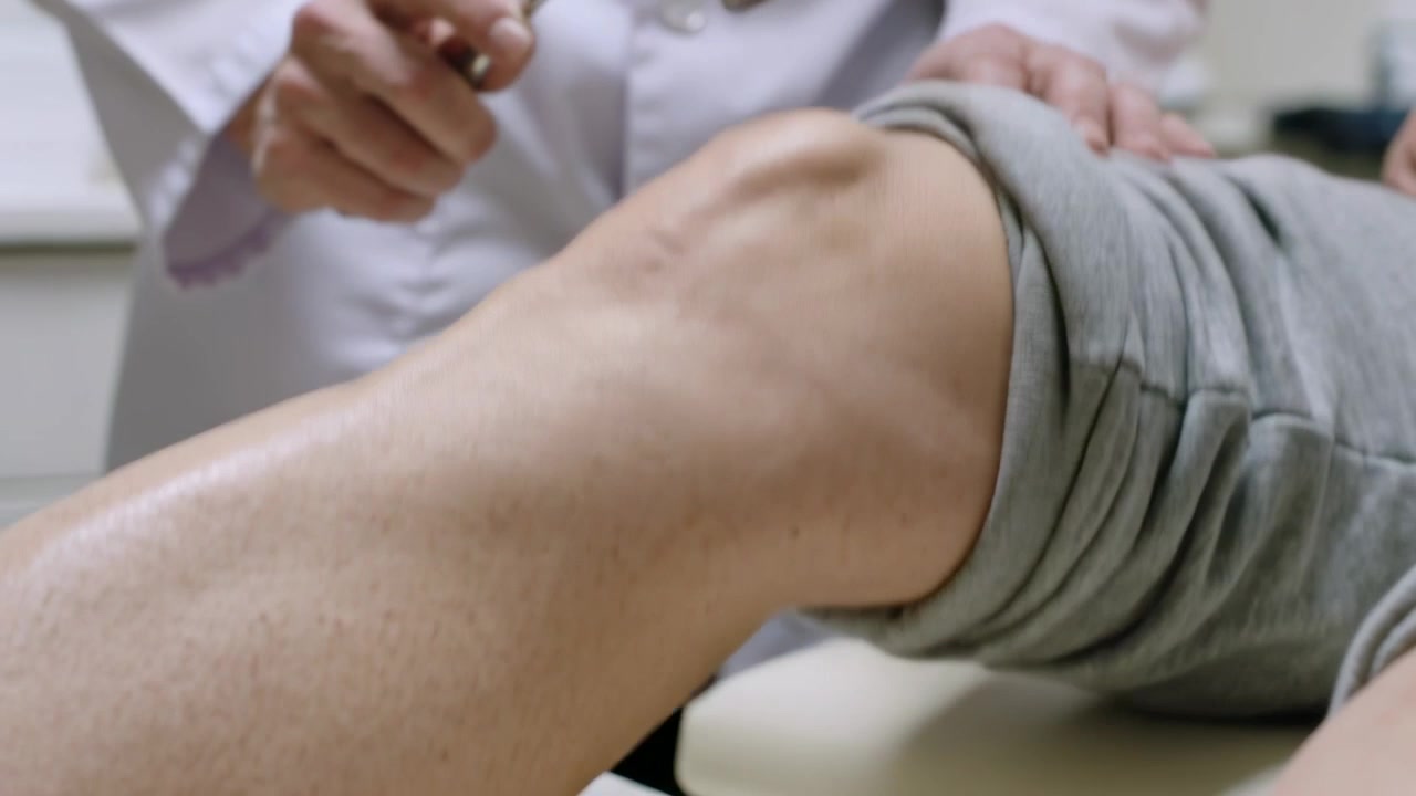 Appoggiando la mano, medico in camice che picchietta il ginocchio destro del paziente al tavolo, testando il riflesso con un martelletto.