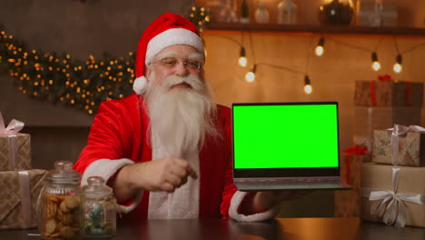 El-Amable-Papá-Noel-Sostiene-Una-Computadora-Portátil-Con-Una-Pantalla-Verde.-Indica-La-Pantalla-Del-Portátil.-Se-Sienta-En-Una-Hermosa-Habitación-Decorada-Para-Una-Feliz-Navidad.-Imágenes-4k-De-Alta-Calidad